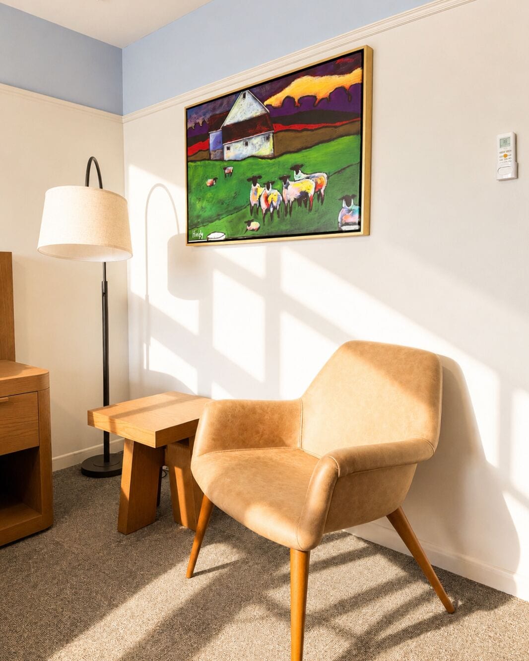 Una habitación con una silla moderna, una mesa de madera, una lámpara de pie y un cuadro con un granero y ovejas en la pared.