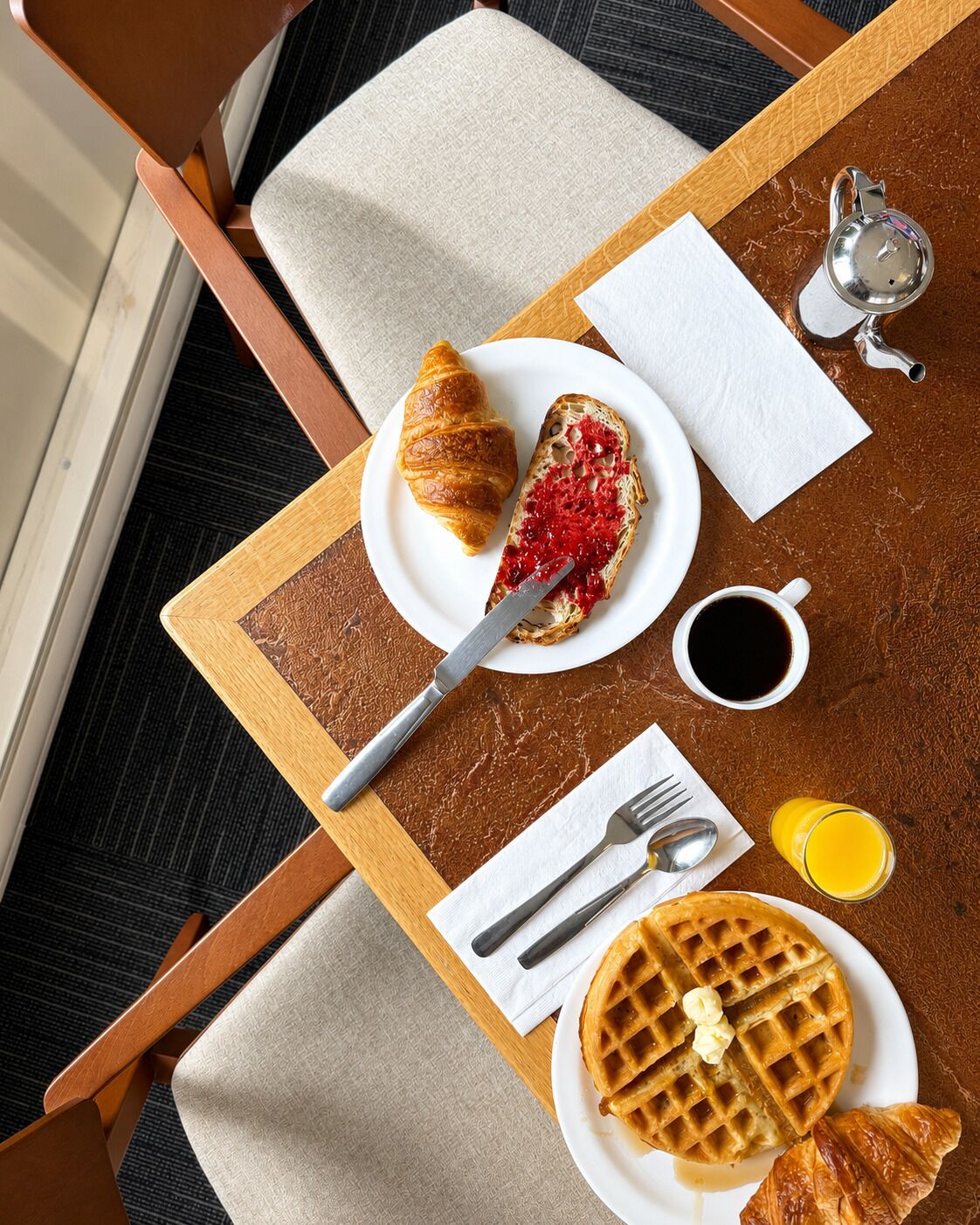 A breakfast tray with croissant, jam, waffle, butter, syrup, coffee, and juice.