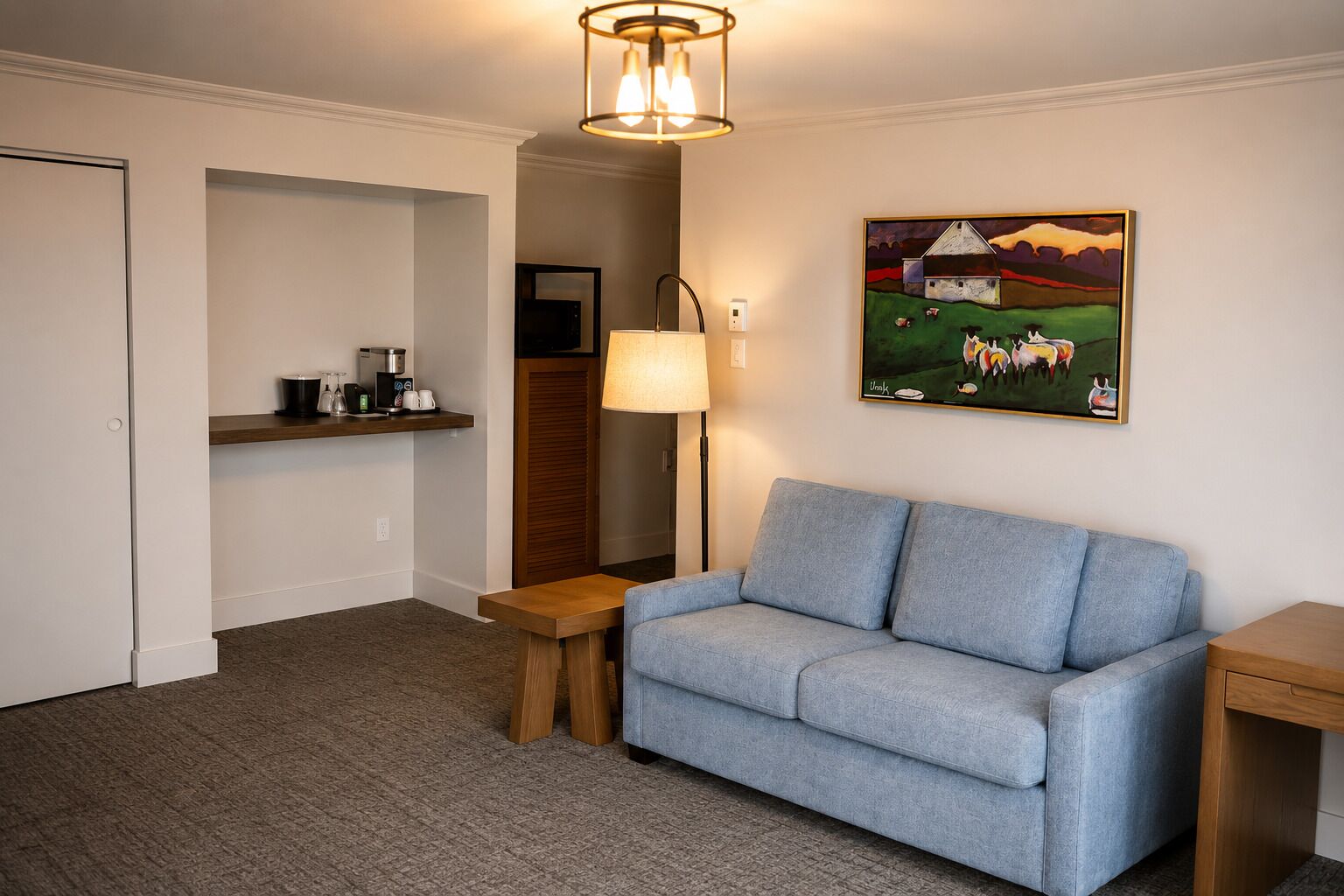 A room with a blue sofa, a wooden side table, a lamp, a painting of a farm scene, and a coffee station.