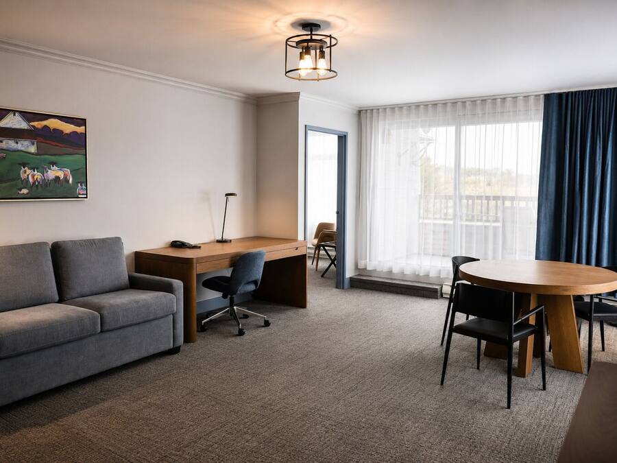 Habitación de hotel con un sofá gris, un escritorio de madera, una mesa redonda y un cuadro en la pared.