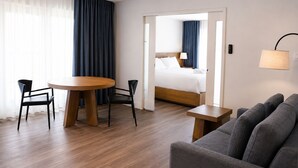 Habitación de hotel con una mesa de comedor de madera, dos sillas, un sofá, una cama con cabecera de madera y una mesita de noche con lámpara.