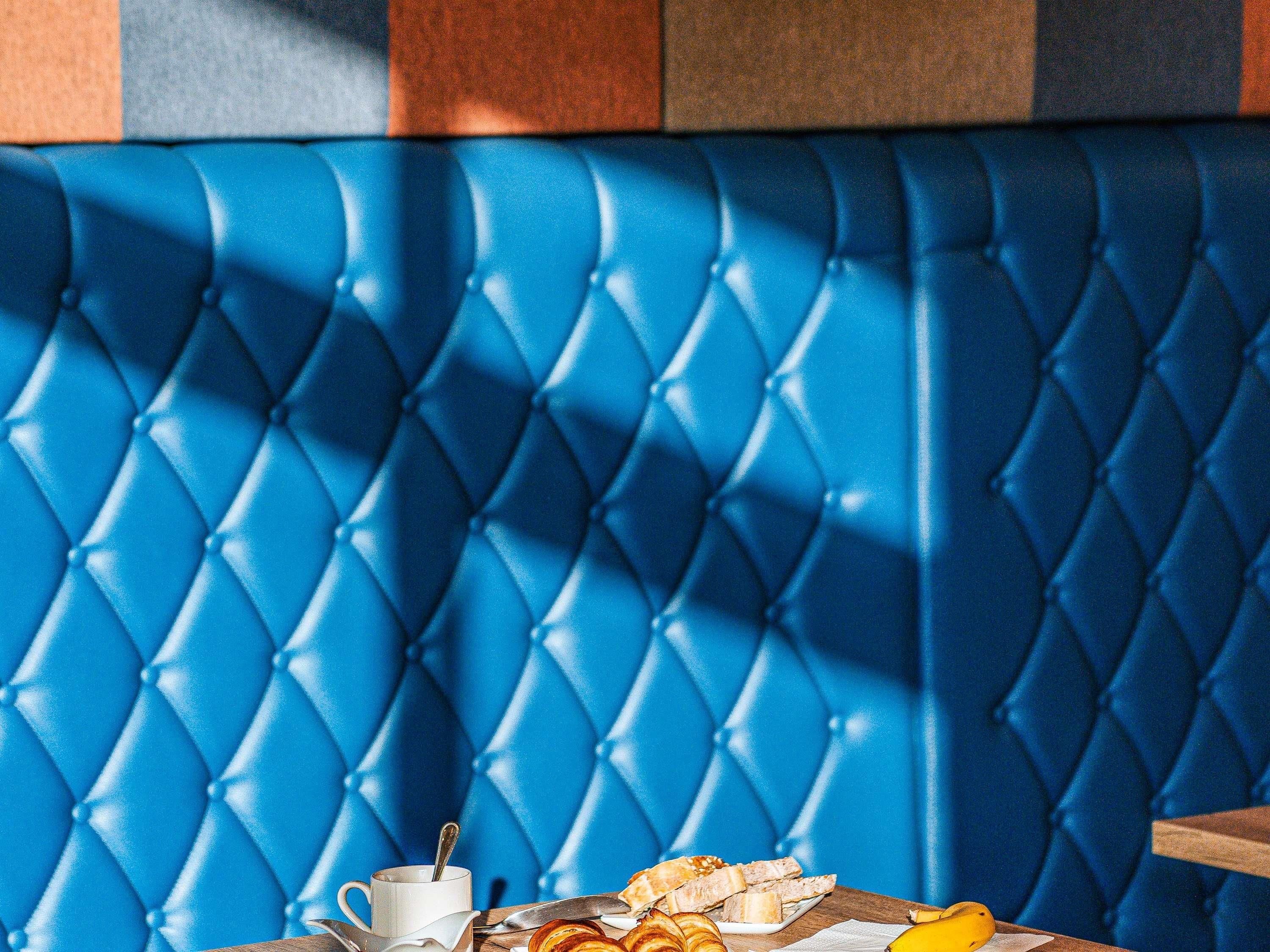 Un banco tapizado azul con diseño capitonado, una taza blanca con cuchara, y un plato con pan y una banana.
