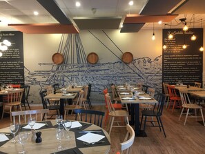 L’intérieur d’un restaurant est décoré de tables et de chaises en bois, d’une fresque murale et de luminaires suspendus.