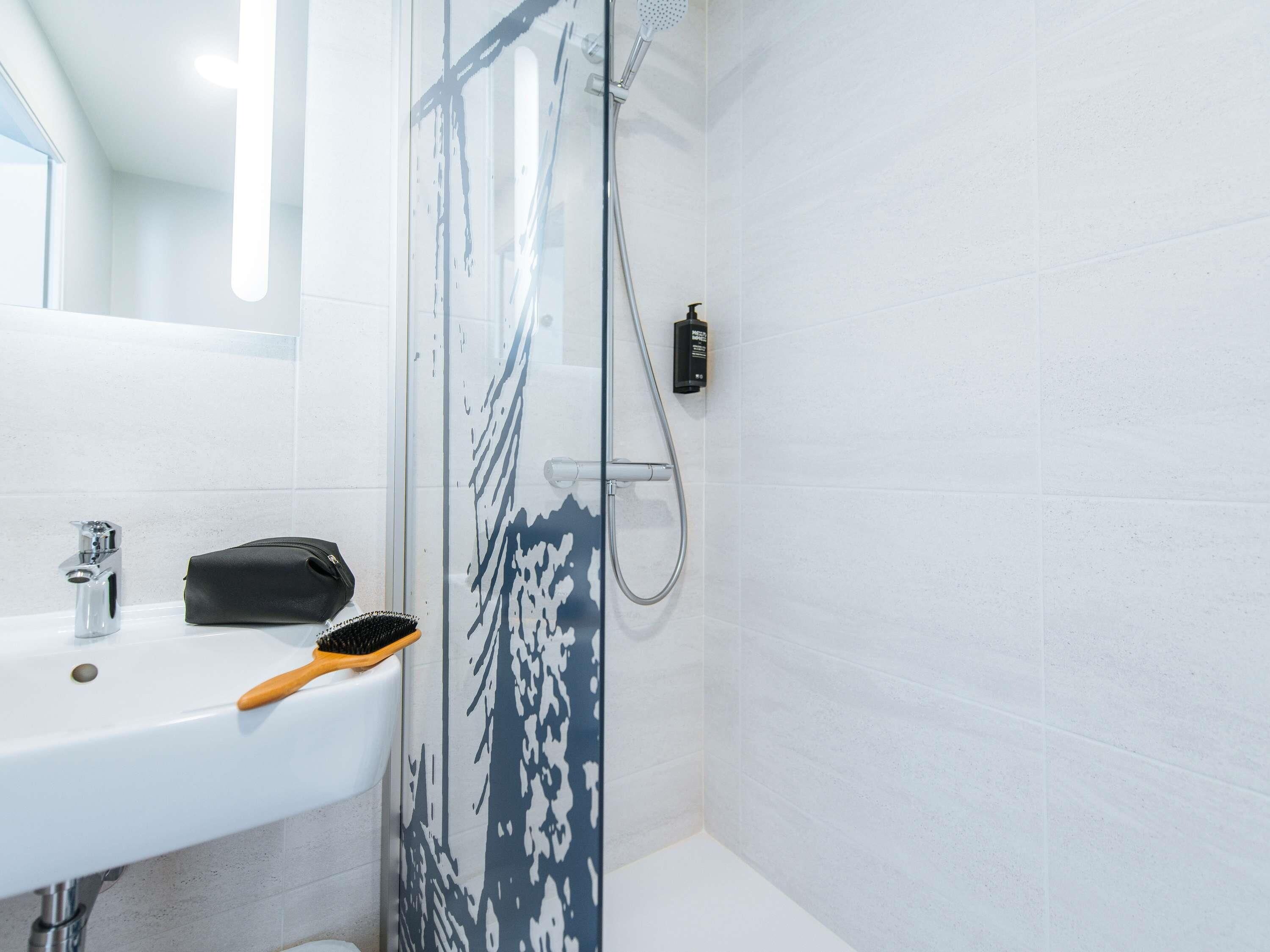 Une salle de bain avec un lavabo blanc, une douchette à main et un sac noir posé sur le comptoir.