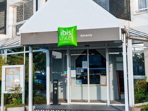 Een hotelentree met een glazen gevel, een groen bord met de tekst ibis Styles Hotels, en een zichtbaar menubord.