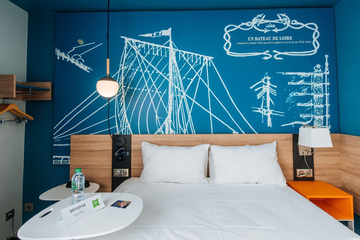 Ein Hotelzimmer mit einem Bett, einem Nachttisch, einem runden Tisch mit einer Flasche und einer Willkommenskarte, sowie einer Wand mit einem Seekarten-Wandgemälde.