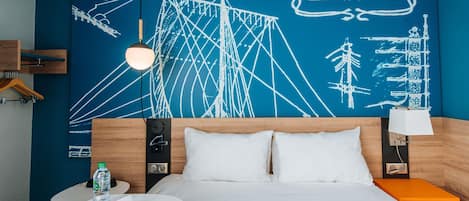 Una habitación de hotel con una cama, una mesita de noche, una mesa redonda con una botella y una tarjeta de bienvenida, y una pared con un mural de temática náutica.