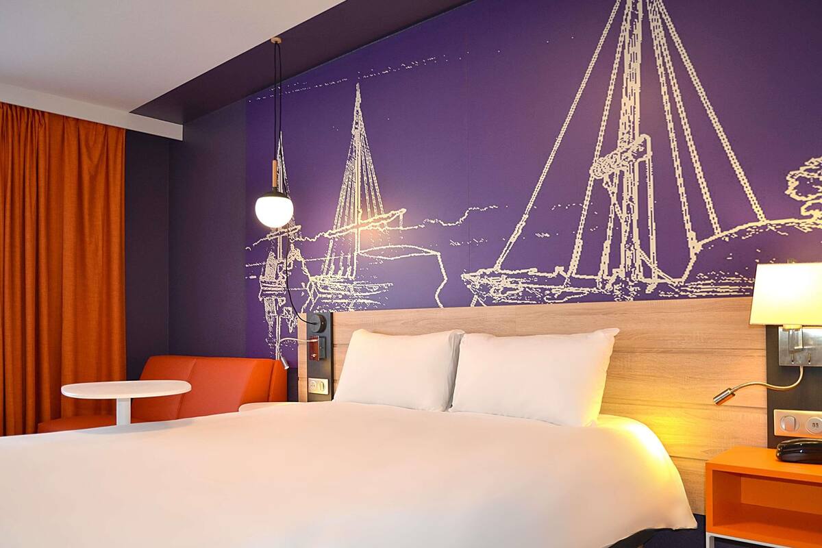 Une chambre d’hôtel avec un lit, une table de chevet, une chaise et une fresque murale représentant des voiliers.