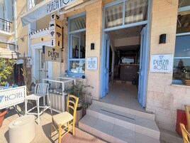Entrada de un hotel con una recepción, una escalera que conduce al interior y mobiliario exterior con sillas y mesas.