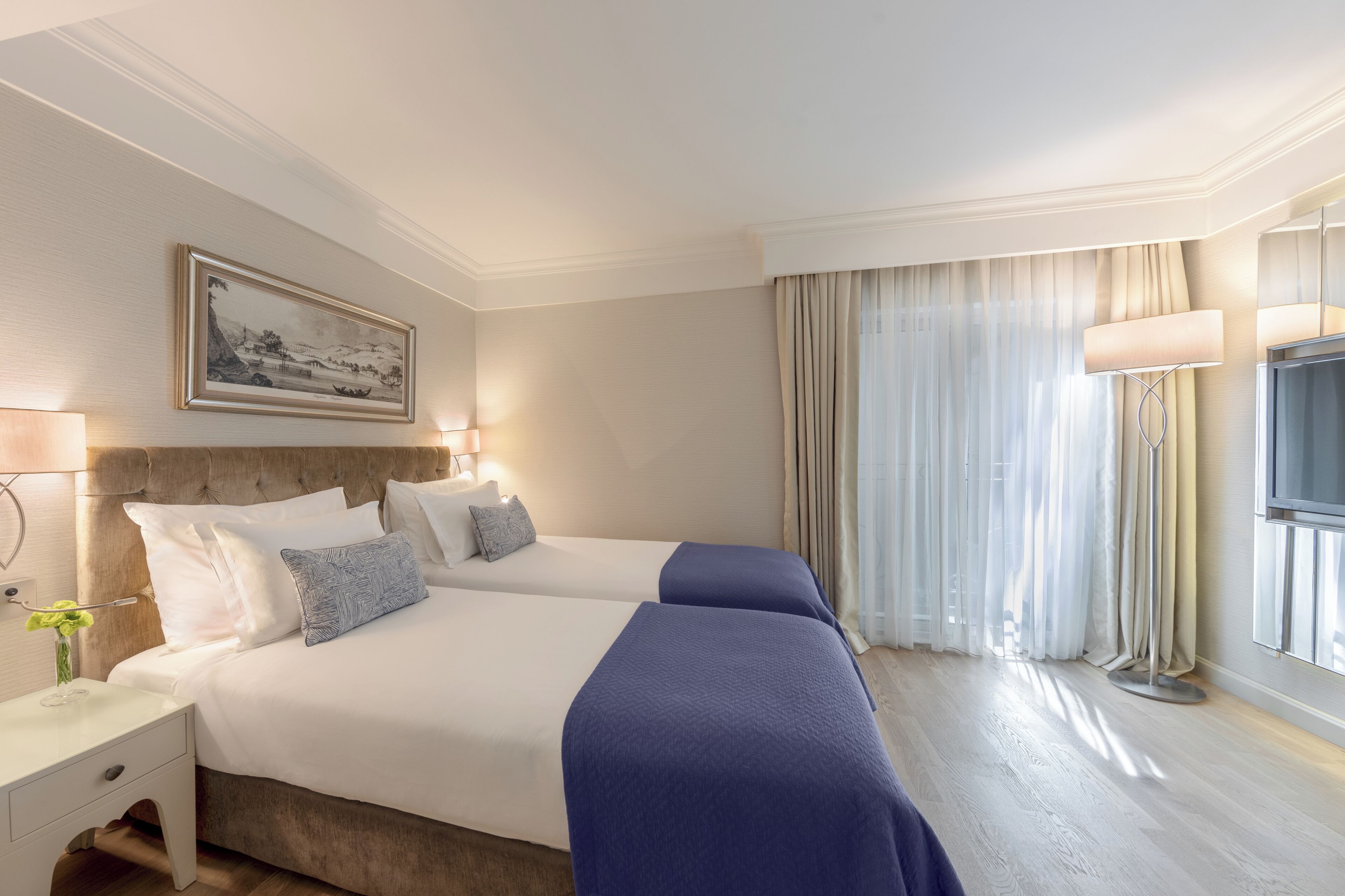 Et hotelværelse med to senge, et natbord, en lampe, et billede og et vindue med gardiner.
