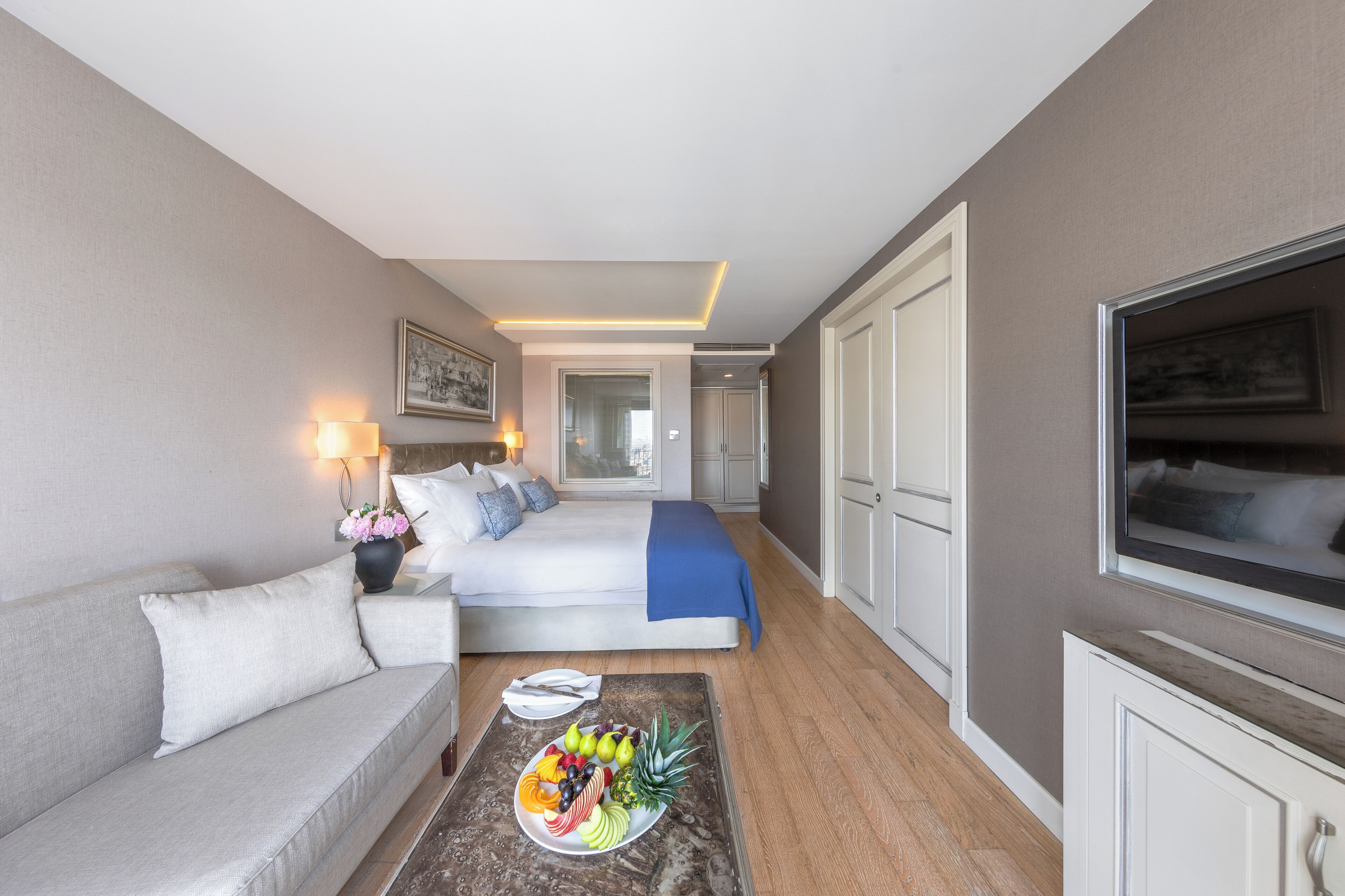 Et hotelværelse med en seng, en sofa, et spisebord med frugt, et fjernsyn og et badeværelse, der kan ses gennem en åben dør.