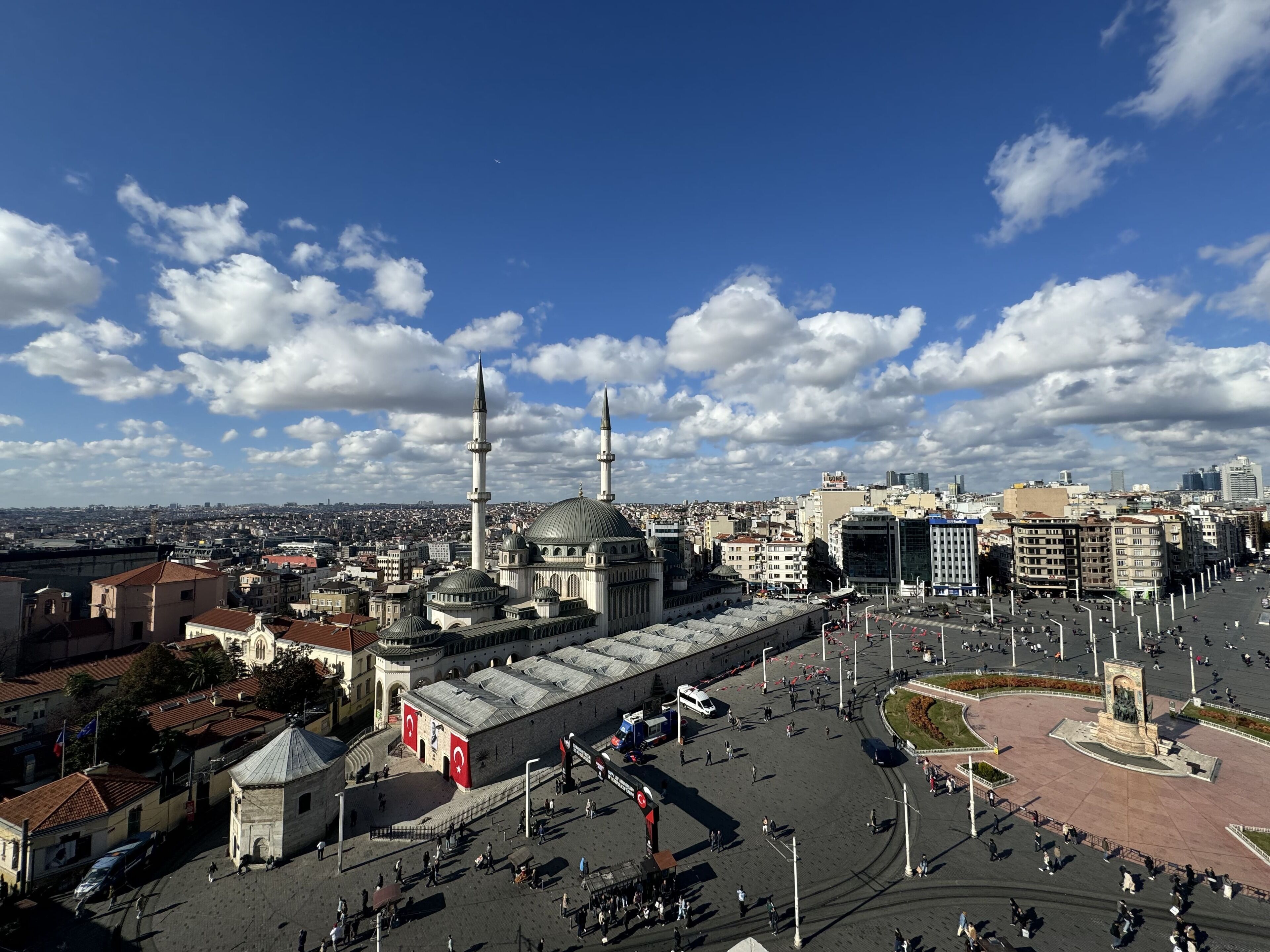 Una amplia plaza abierta con una mezquita destacada que cuenta con dos altos minaretes y una cúpula central, rodeada de edificios modernos y un cielo despejado.