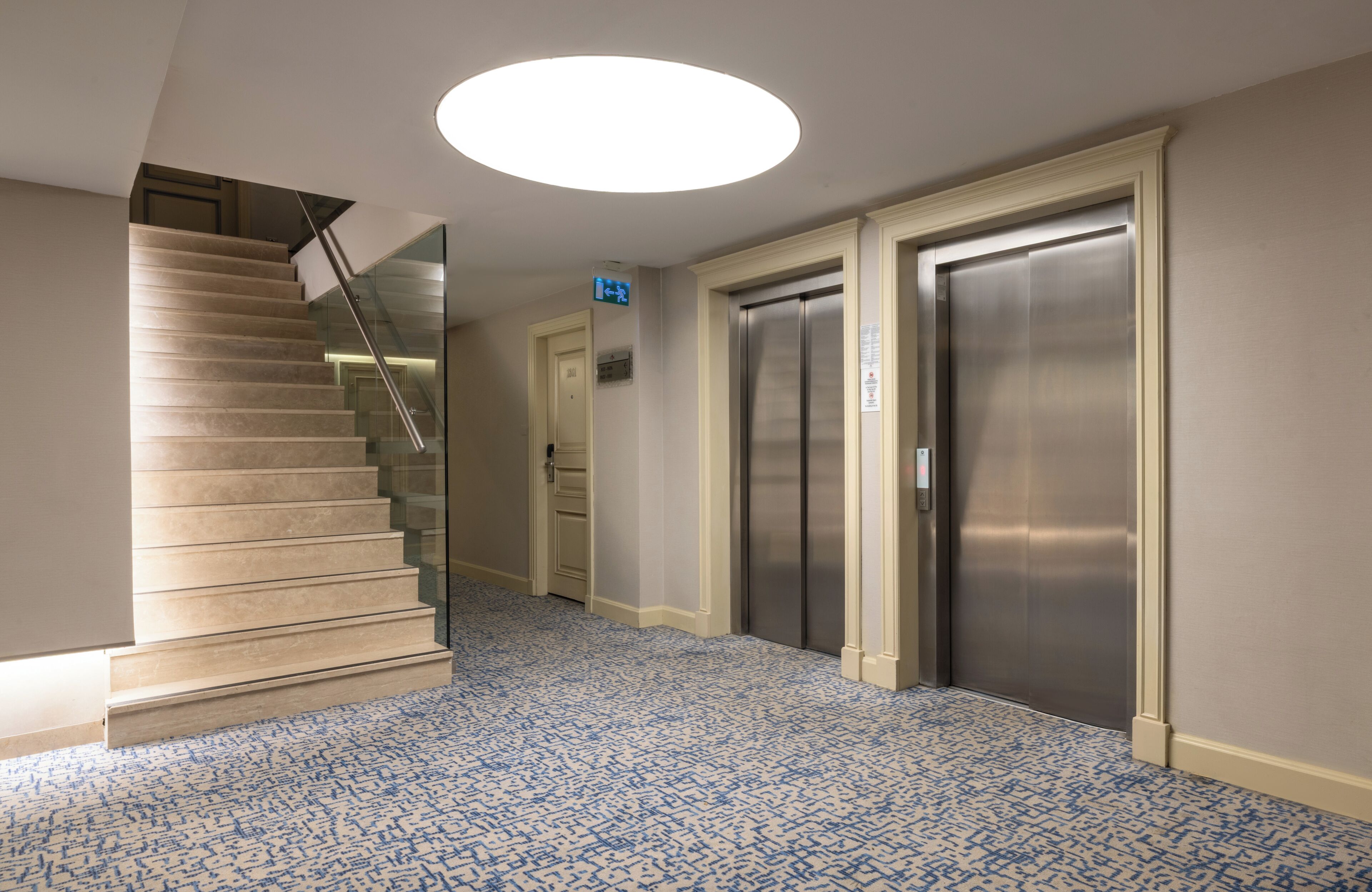 A modern interior with a carpeted staircase, two elevators, and a recessed ceiling light.