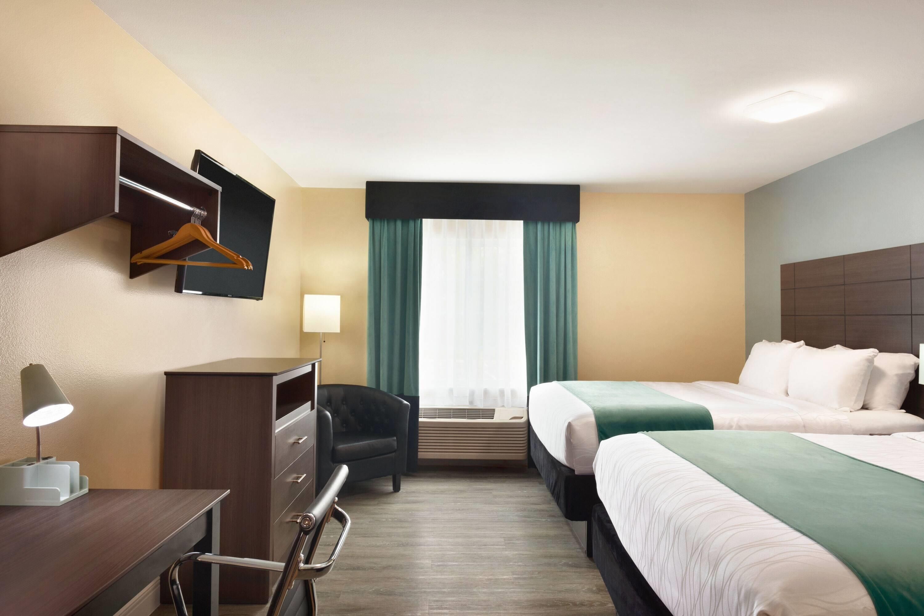 Ein Hotelzimmer mit zwei Betten, einem Schreibtisch mit Lampe, einem Stuhl, einem Kleiderschrank und einem Fenster mit Vorhängen.