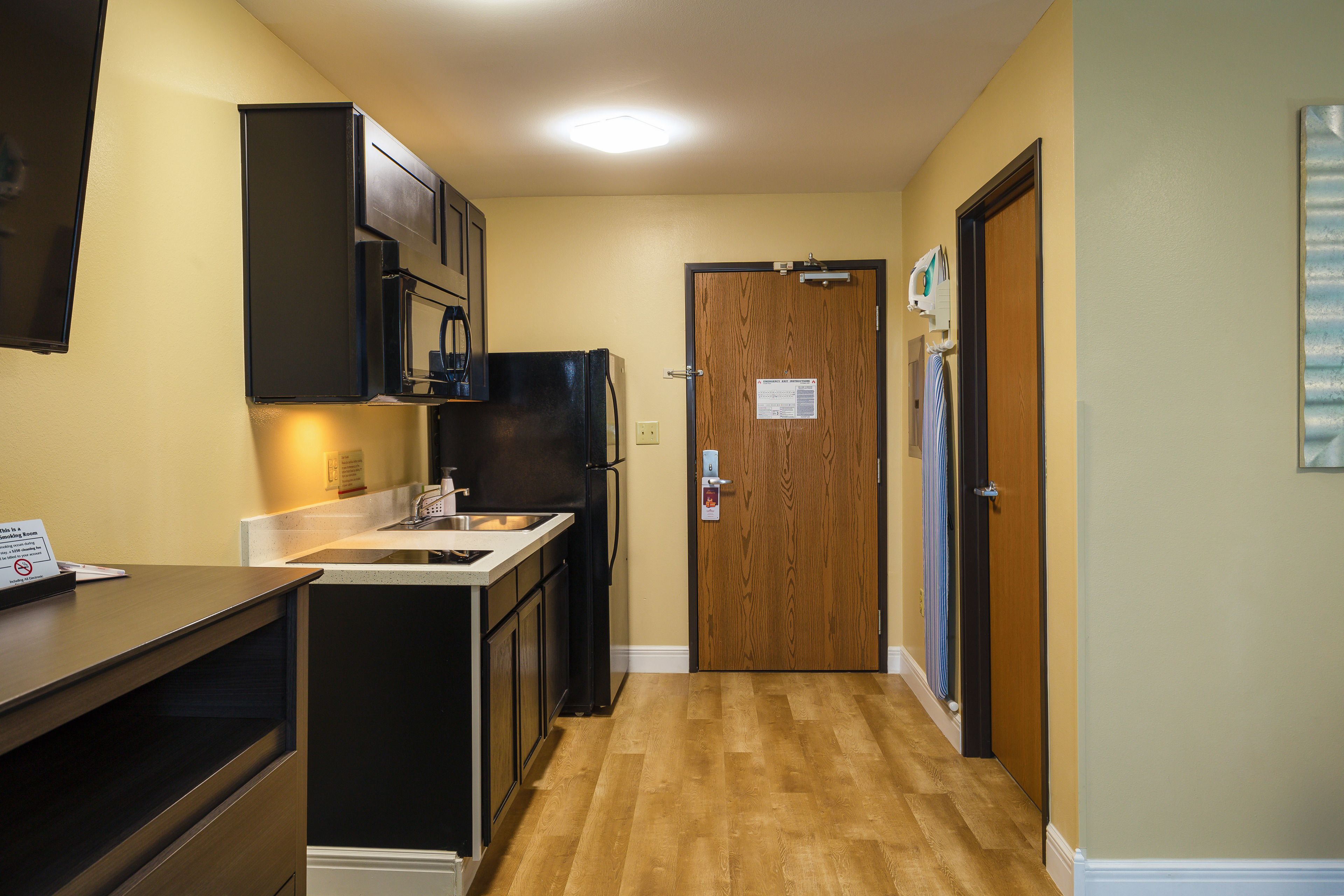 Un pasillo estrecho con una zona de cocina a la izquierda, que incluye un refrigerador, estufa y armarios. Al final del pasillo hay una puerta de madera con un letrero.