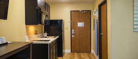 A narrow hallway with a kitchen area on the left, featuring a refrigerator, stove, and cabinets. A wooden door with a sign is at the end of the hallway.