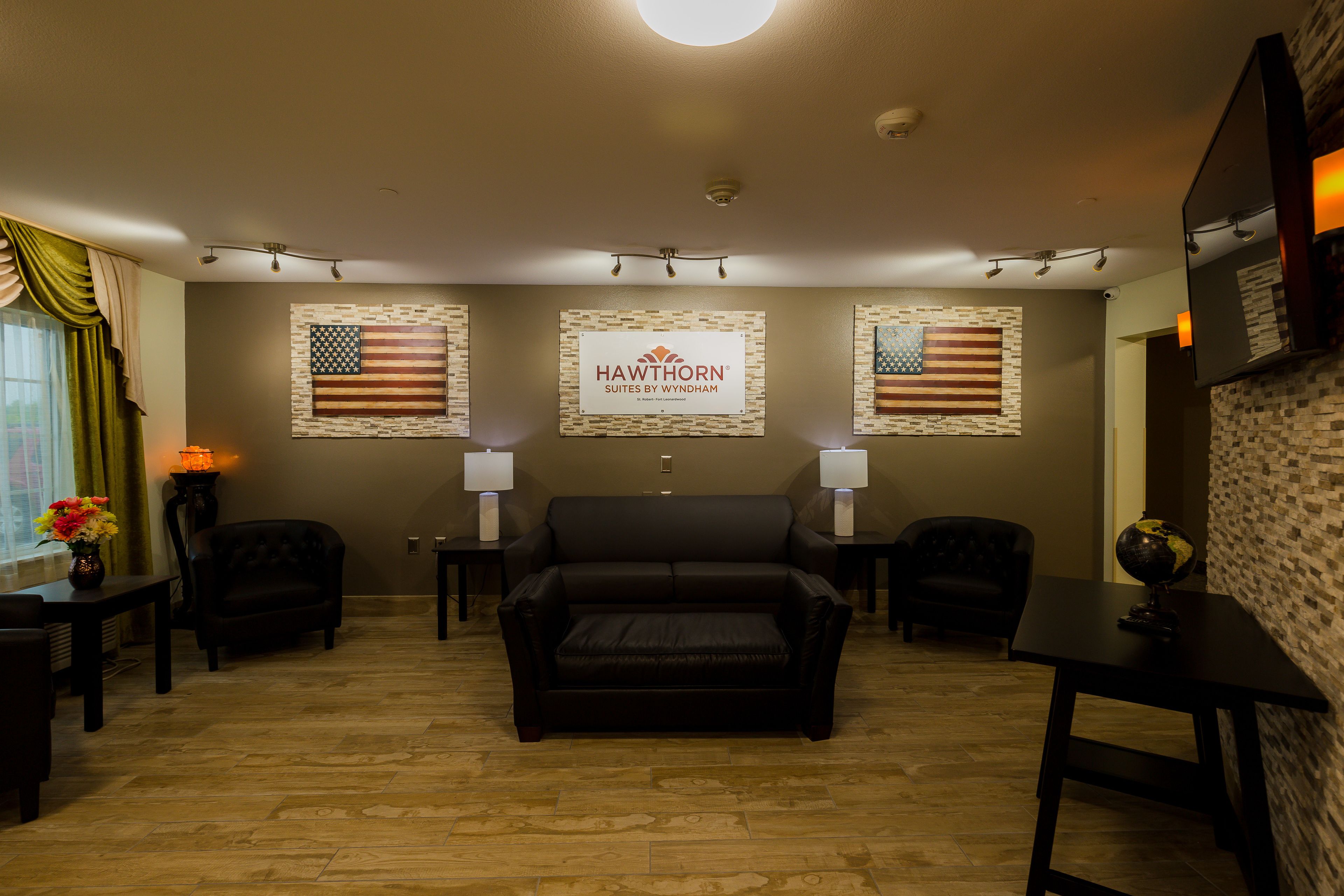 Quarto de hotel com sofá de couro, duas poltronas, uma mesinha e duas luminárias. Há bandeiras americanas nas paredes e uma placa da Hawthorn Suites by Wyndham.