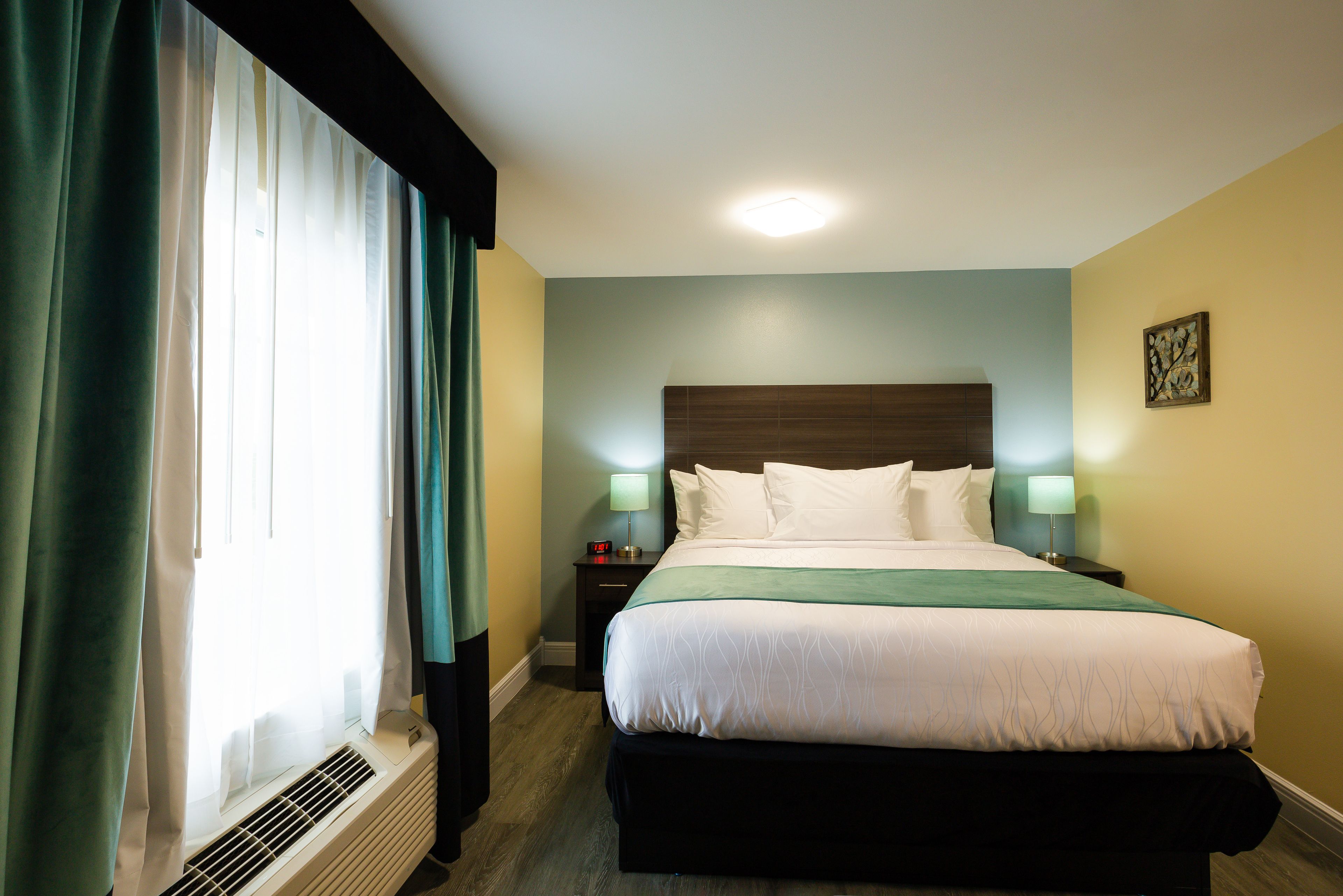 Een hotelkamer met een groot bed, een nachtkastje met een lamp, een raam met gordijnen en een aan de muur hangend schilderij.