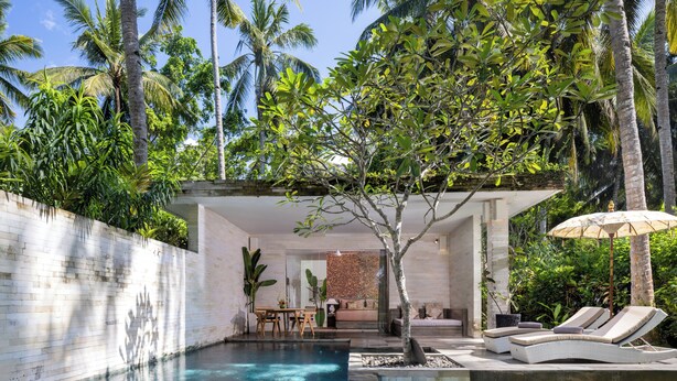 Un área de piscina moderna con una tumbona, una mesa y un espacio de estar techado, rodeado de exuberante vegetación y palmeras.