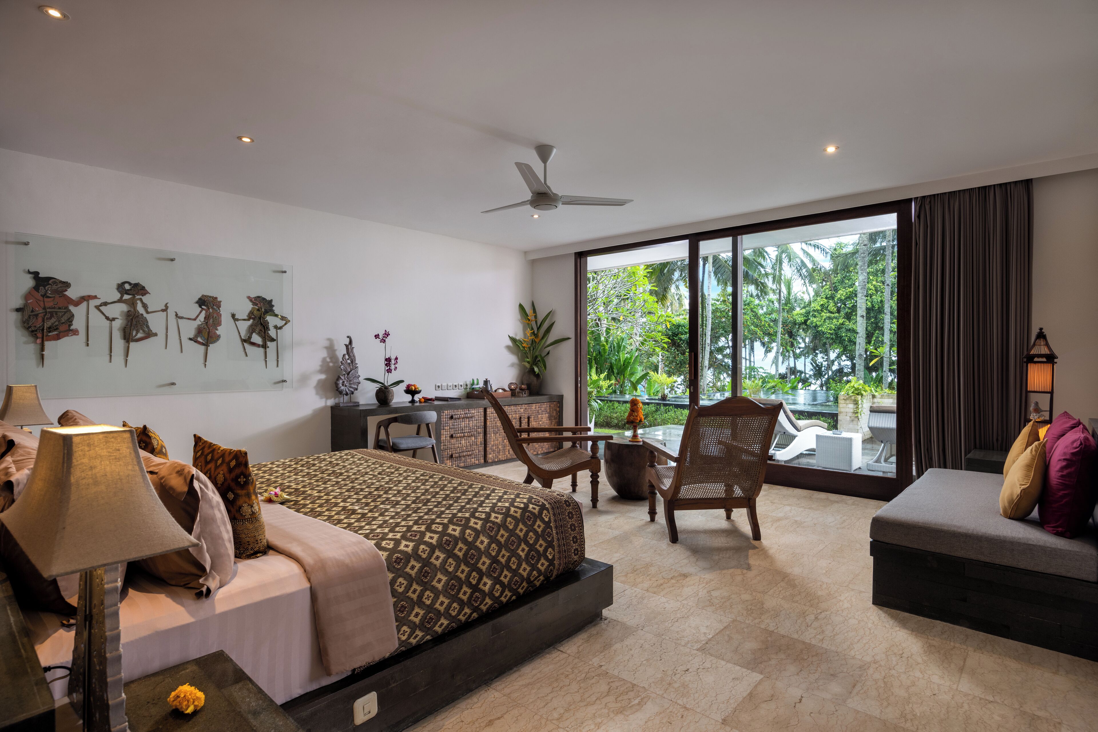 A modern bedroom with a bed, desk, chairs, and a balcony with a view of greenery.