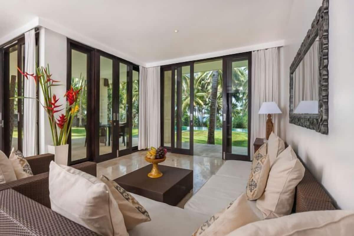 A modern living room with a sofa, a coffee table with fruit, and a view of a tropical garden through glass doors.