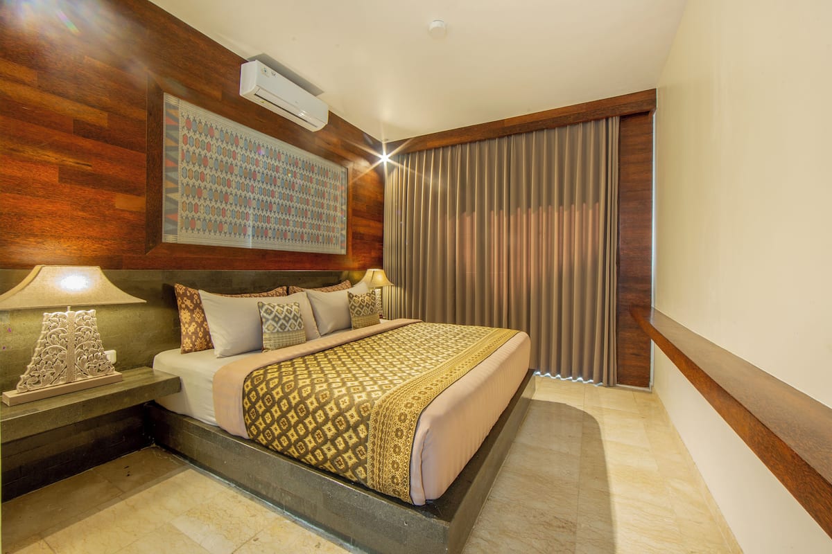 A hotel room with a bed, nightstands, a wall-mounted air conditioner, and a large window with curtains.