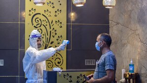 Una persona con equipo de protección usando un termómetro infrarrojo en otra persona, en una zona de recepción con un muro decorativo y un pequeño taburete con dispensador de gel hidroalcohólico.