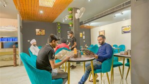 Un comedor moderno con sillas color turquesa y patas doradas, una mesa redonda con un menú, y un techo con paneles de madera y plantas colgantes.