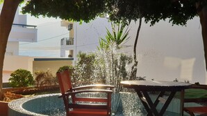 A circular water feature with chairs and a table, surrounded by greenery and a building in the background.
