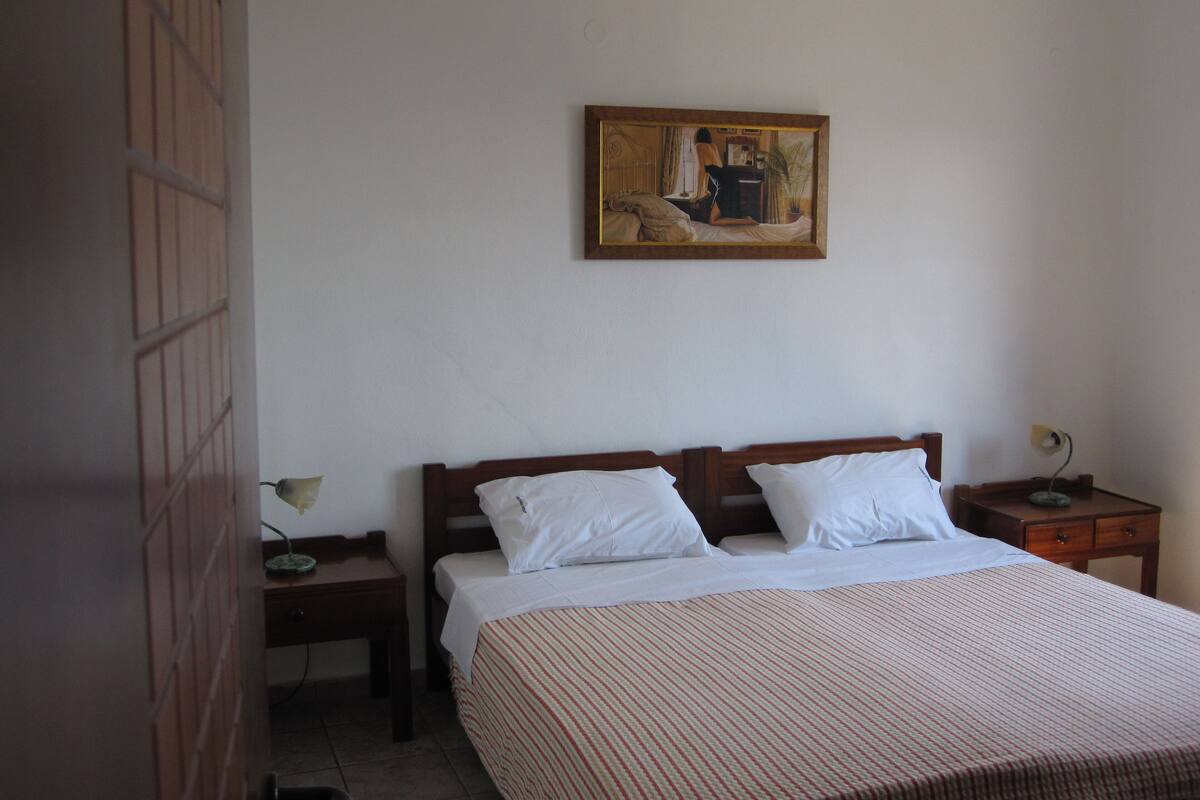 Un lit bien fait, avec du linge de lit blanc, une tête de lit en bois et une couvre-lit à rayures. Un tableau est accroché au mur au-dessus du lit, ainsi qu’une lampe de chevet et une petite table avec une lampe placée à côté du lit.