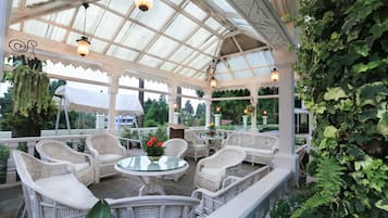 A covered patio area with wicker furniture and a glass table, surrounded by greenery and a white fence.