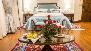 Quarto de hotel com cama, mesa de vidro com frutas, vaso de flores e tapete decorativo.
