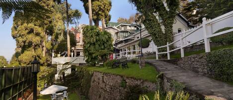 A lush garden with a stone pathway, white outdoor furniture, and a white fence leading to a building with a green roof.