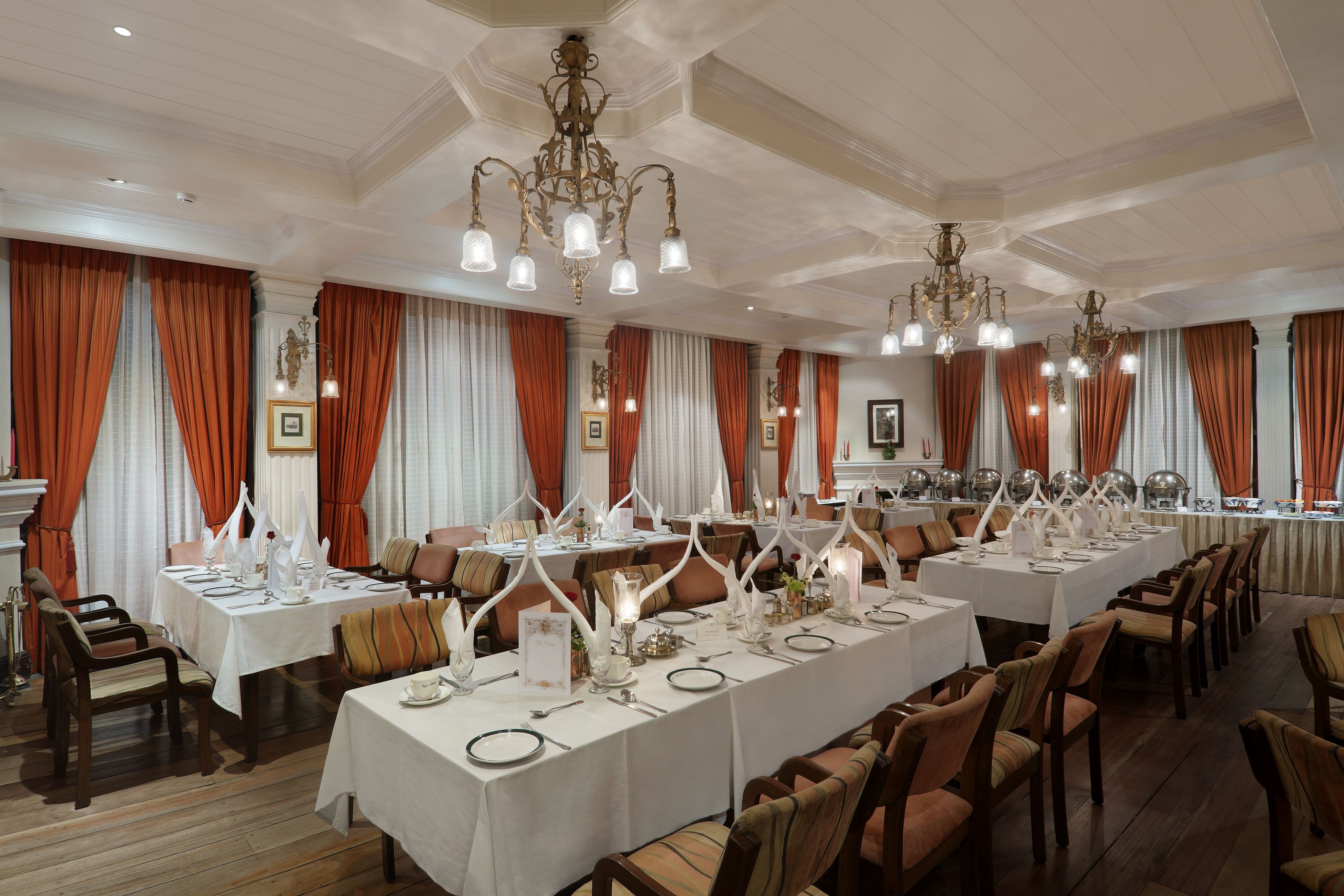 A formal dining room with multiple tables set for service, elegant chandeliers, and classic drapery.
