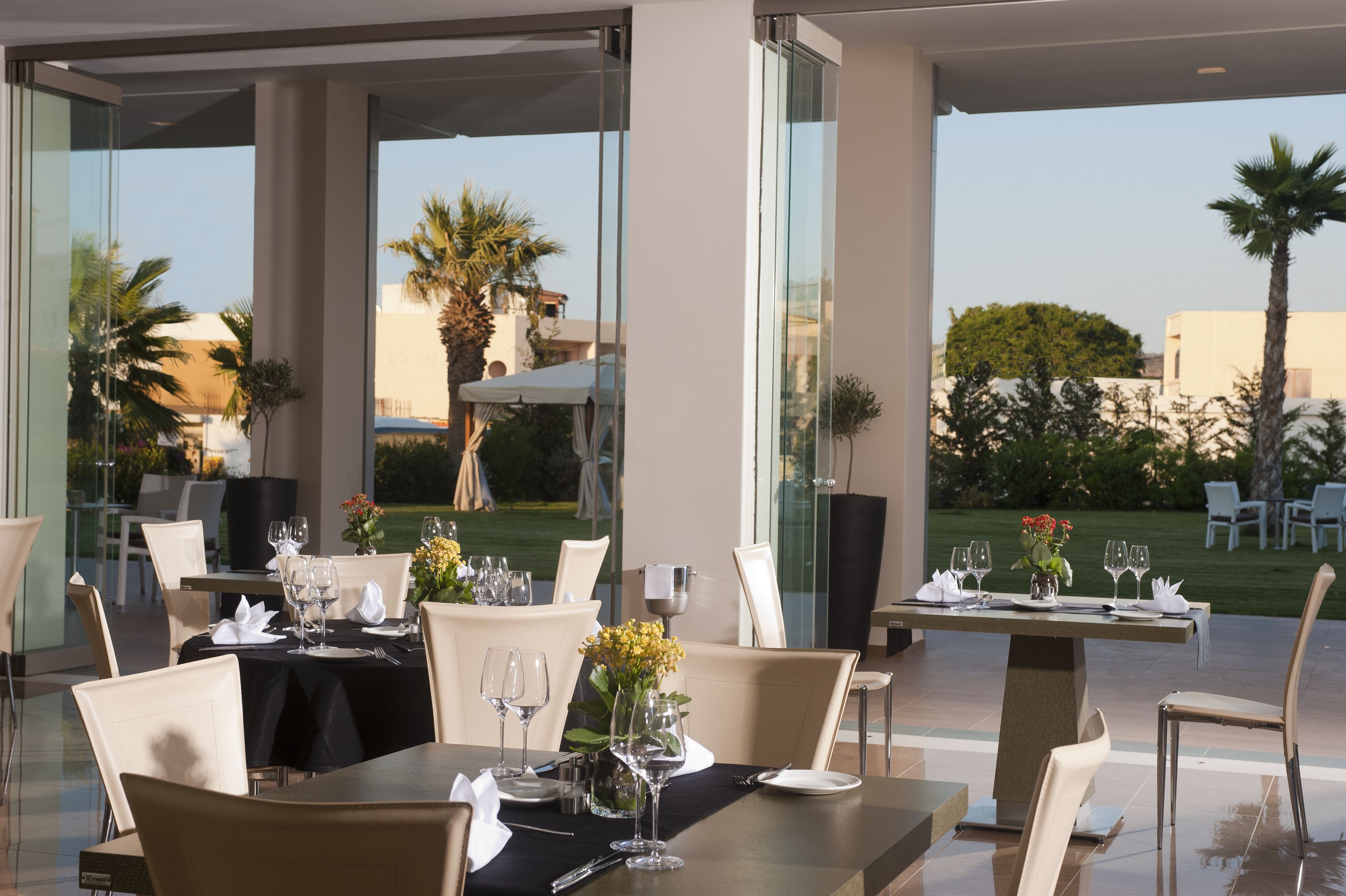 Un área de comedor al aire libre con mesas preparadas para dos, con divisiones de vidrio y vistas al jardín y edificios al fondo.