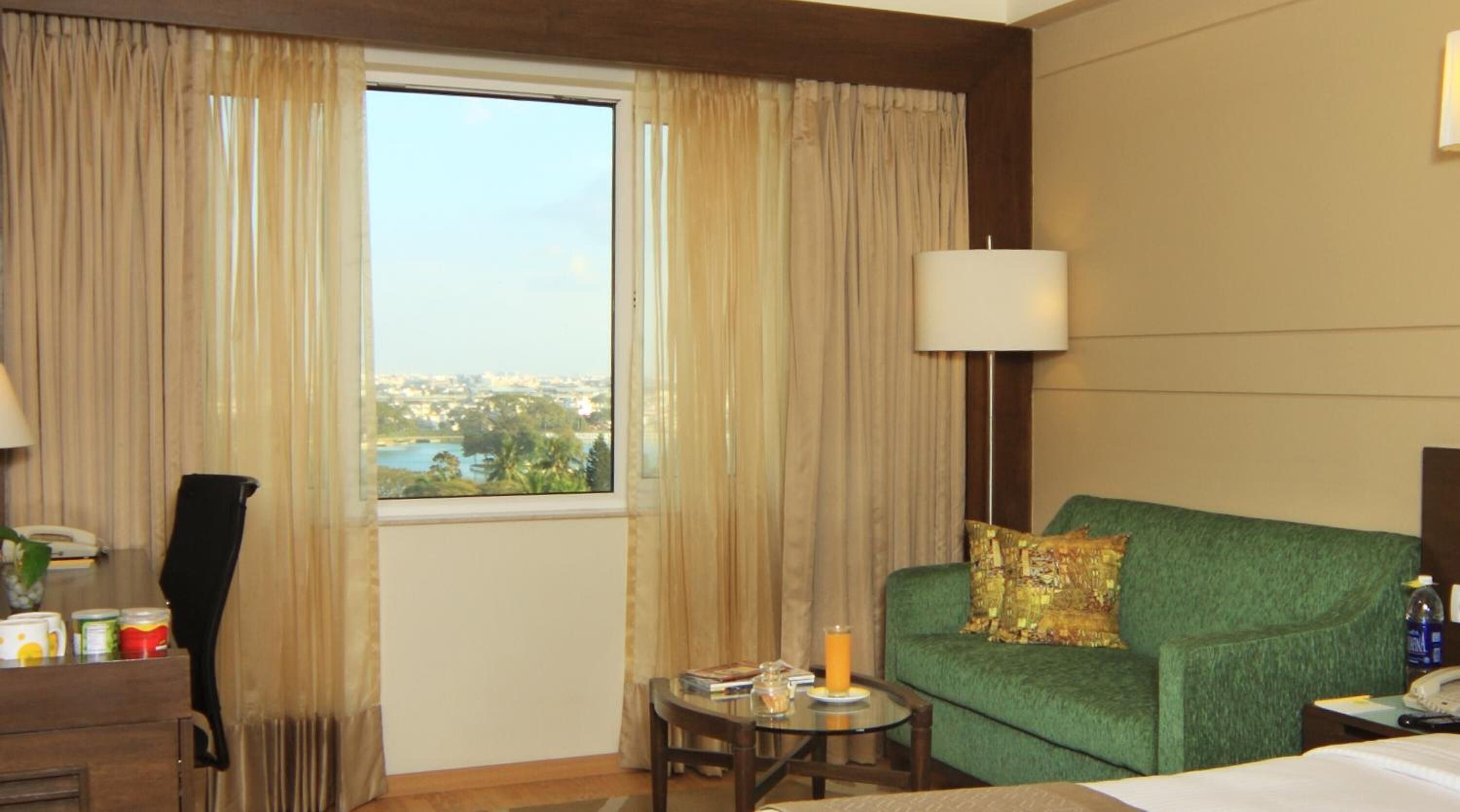 A hotel room with a green sofa, a small table with a lamp, a window with curtains, and a view of a cityscape.