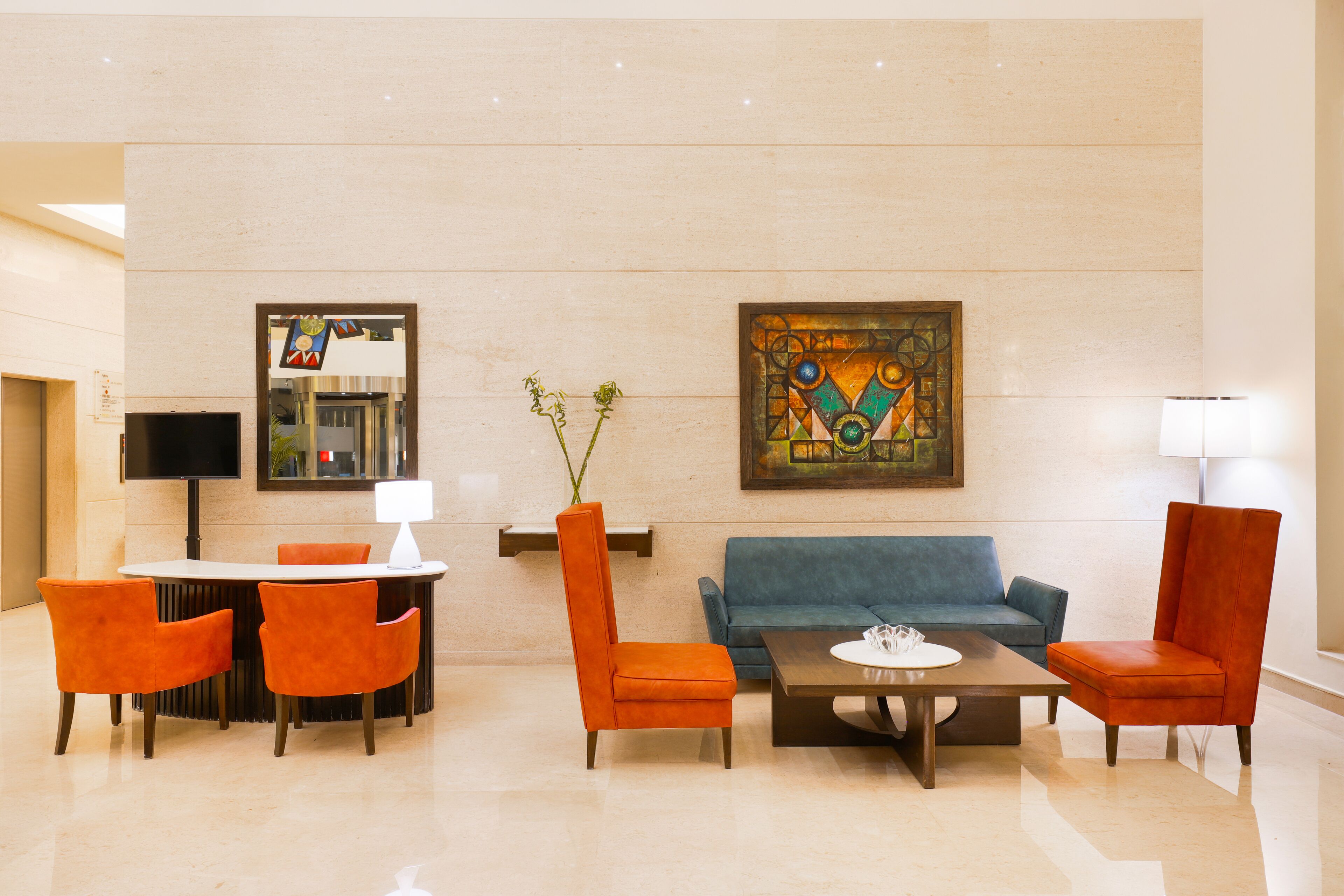 A lobby with a round table, orange chairs, a blue sofa, and a painting on the wall.