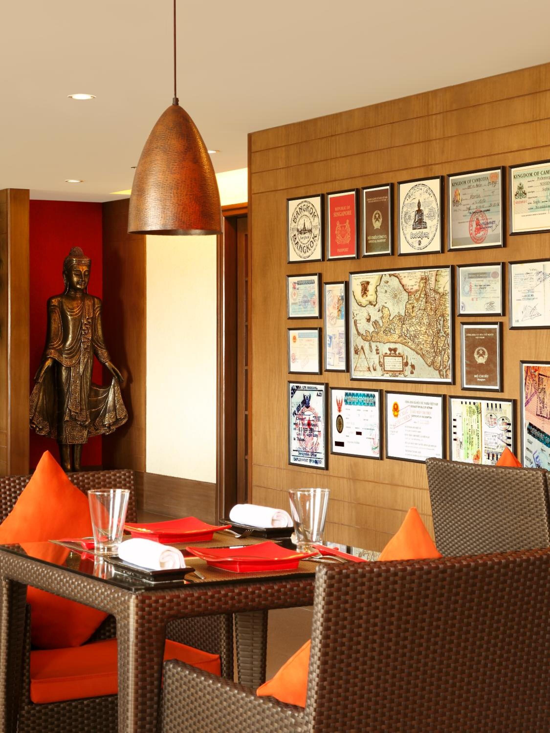 A dining area with wicker chairs and orange cushions, a Buddha statue, and a wall decorated with various certificates and maps.