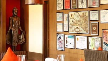 A dining area with wicker chairs and orange cushions, a Buddha statue, and a wall decorated with various certificates and maps.
