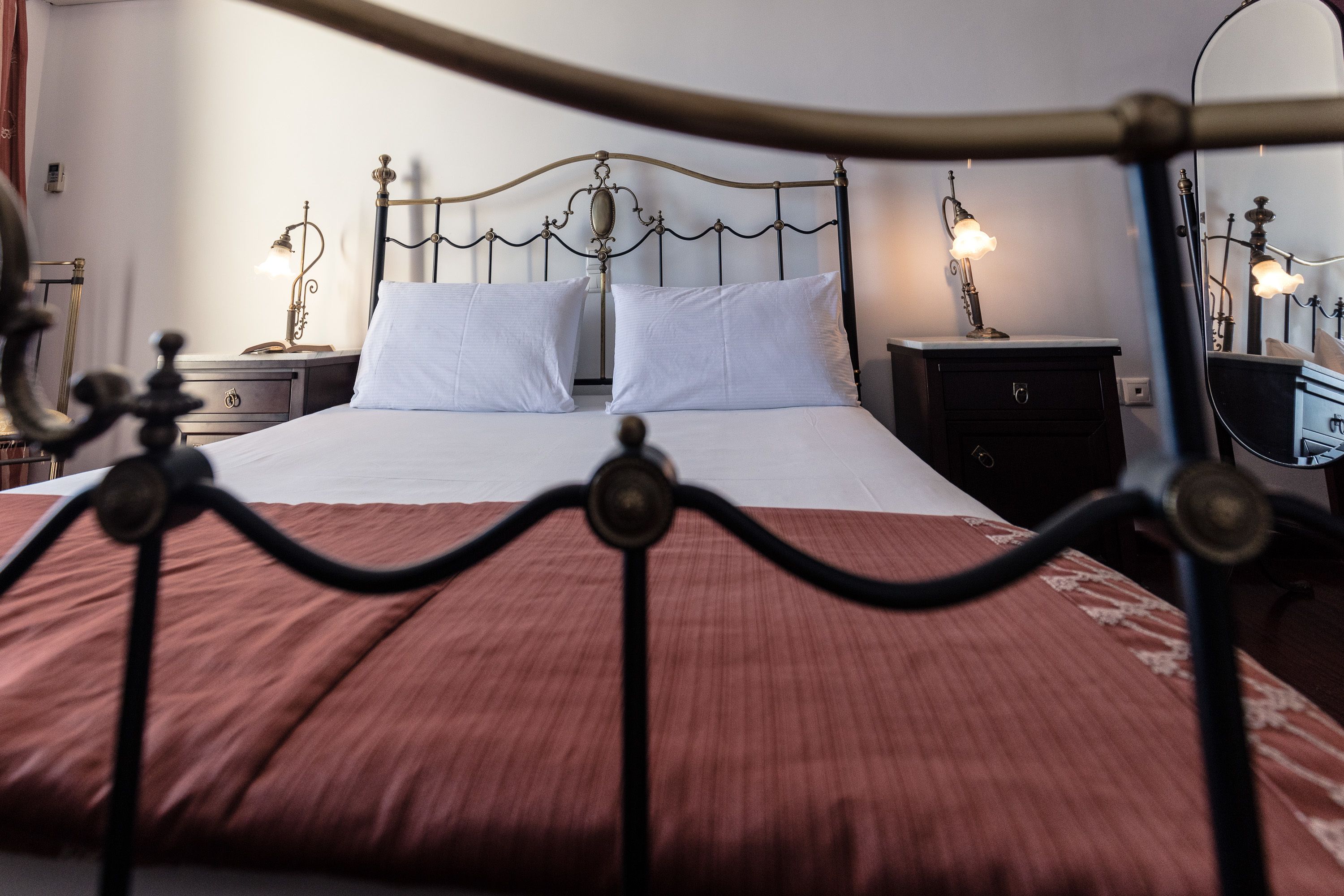 Una cama bien hecha con cabecera de metal, dos mesitas de noche con lámparas y un espejo en la pared.