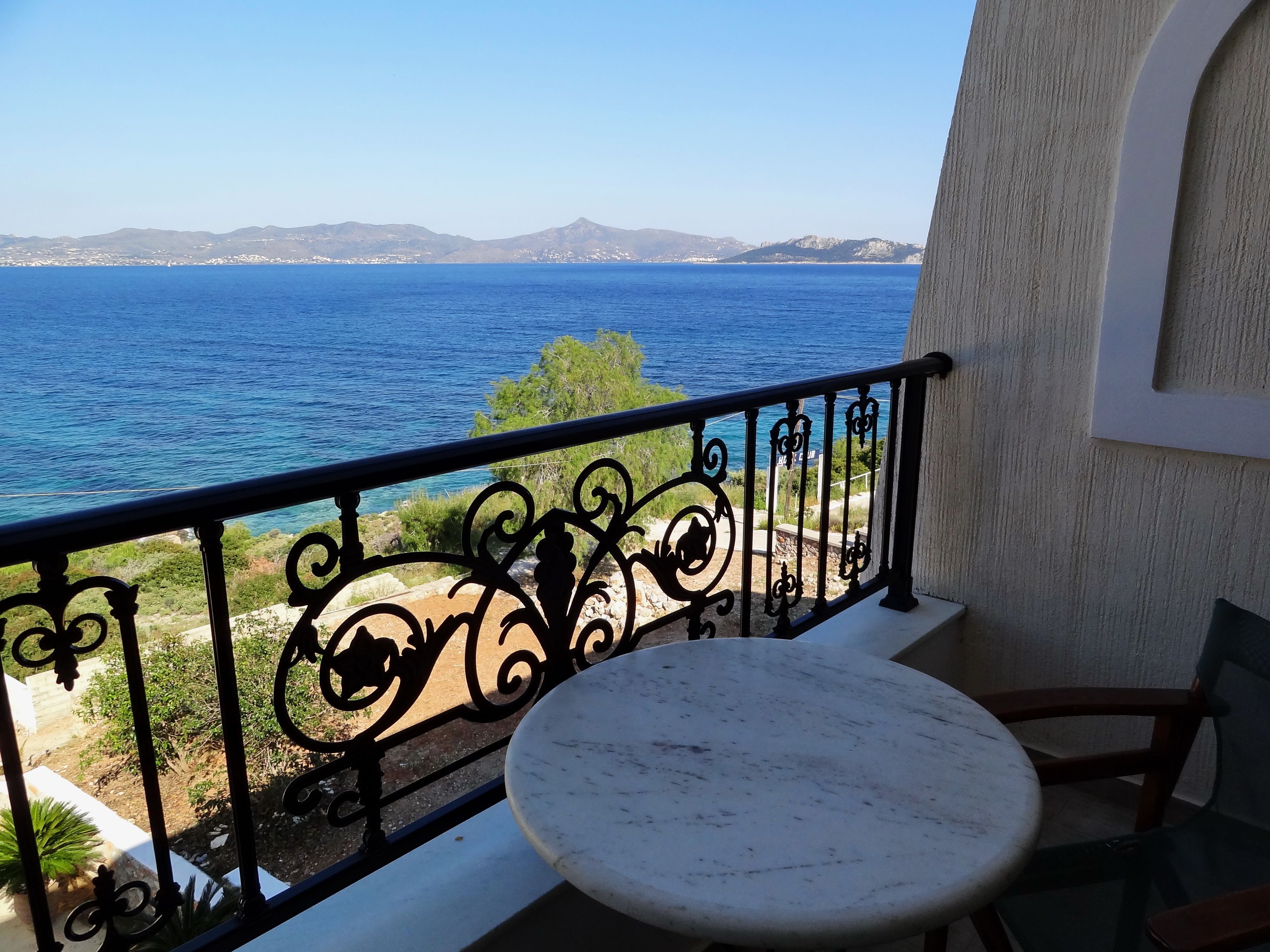 A balcony with a round table and a chair, overlooking a coastal view with mountains in the distance.