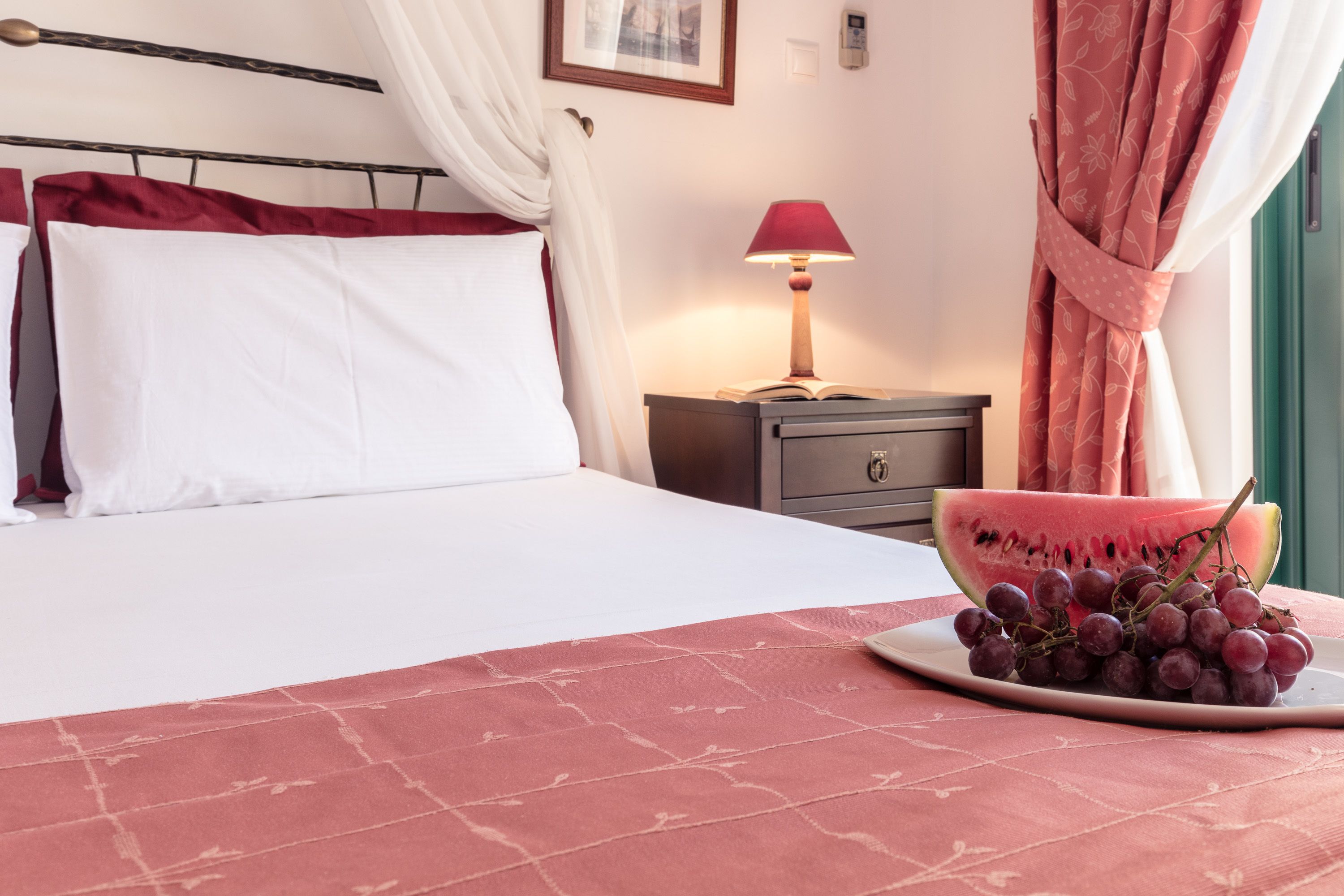 A neatly made bed with white linens, a bedside table with a lamp, a bowl of grapes, and a slice of watermelon on the bed.
