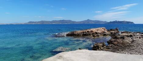 Vista costera con aguas azul cristalino, costa rocosa y montañas al fondo.