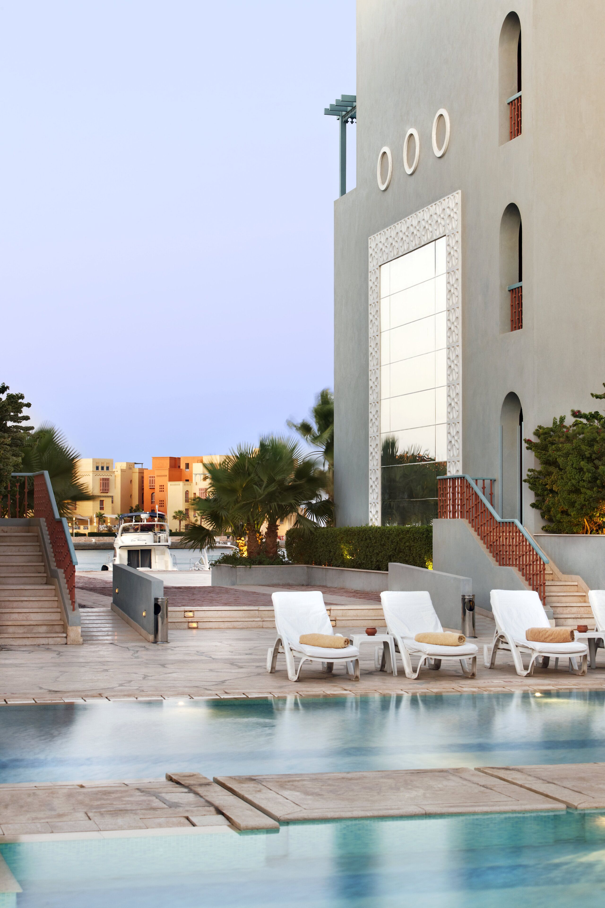 Zona de piscina con sillones reclinables y una escalera que conduce a un edificio con un ventanal grande y tres ventanas circulares.