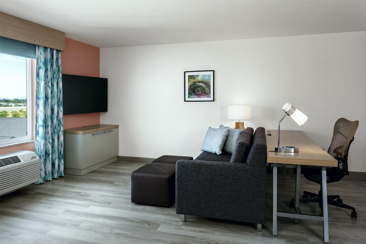 A modern hotel room with a sofa, a desk, a lamp, a TV, and a window with curtains.