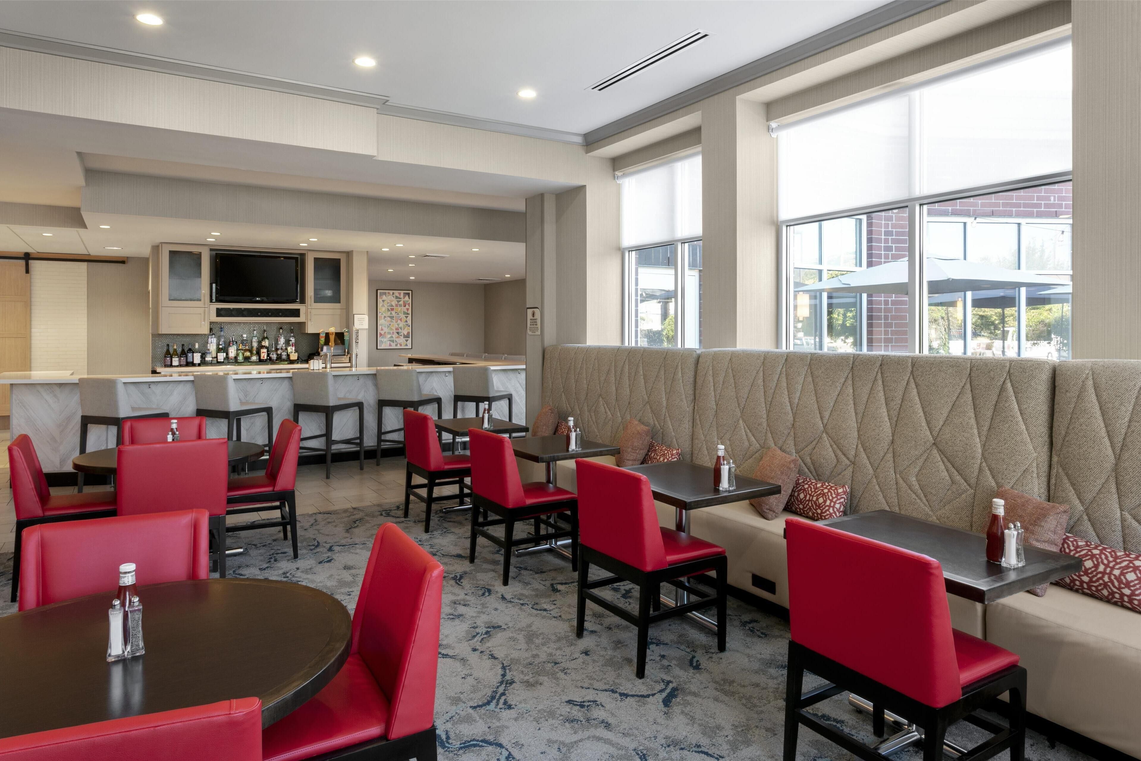 Un espace repas moderne avec des chaises et des tables rouges, un bar avec des bouteilles exposées et de grandes fenêtres offrant une vue sur l’extérieur.
