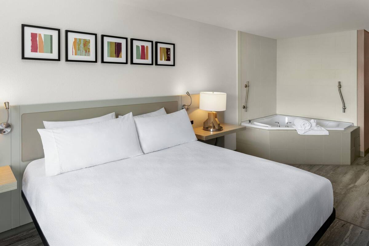 A hotel room with a large bed, a bedside table with a lamp, a small table with a towel, and a bathtub with a showerhead.