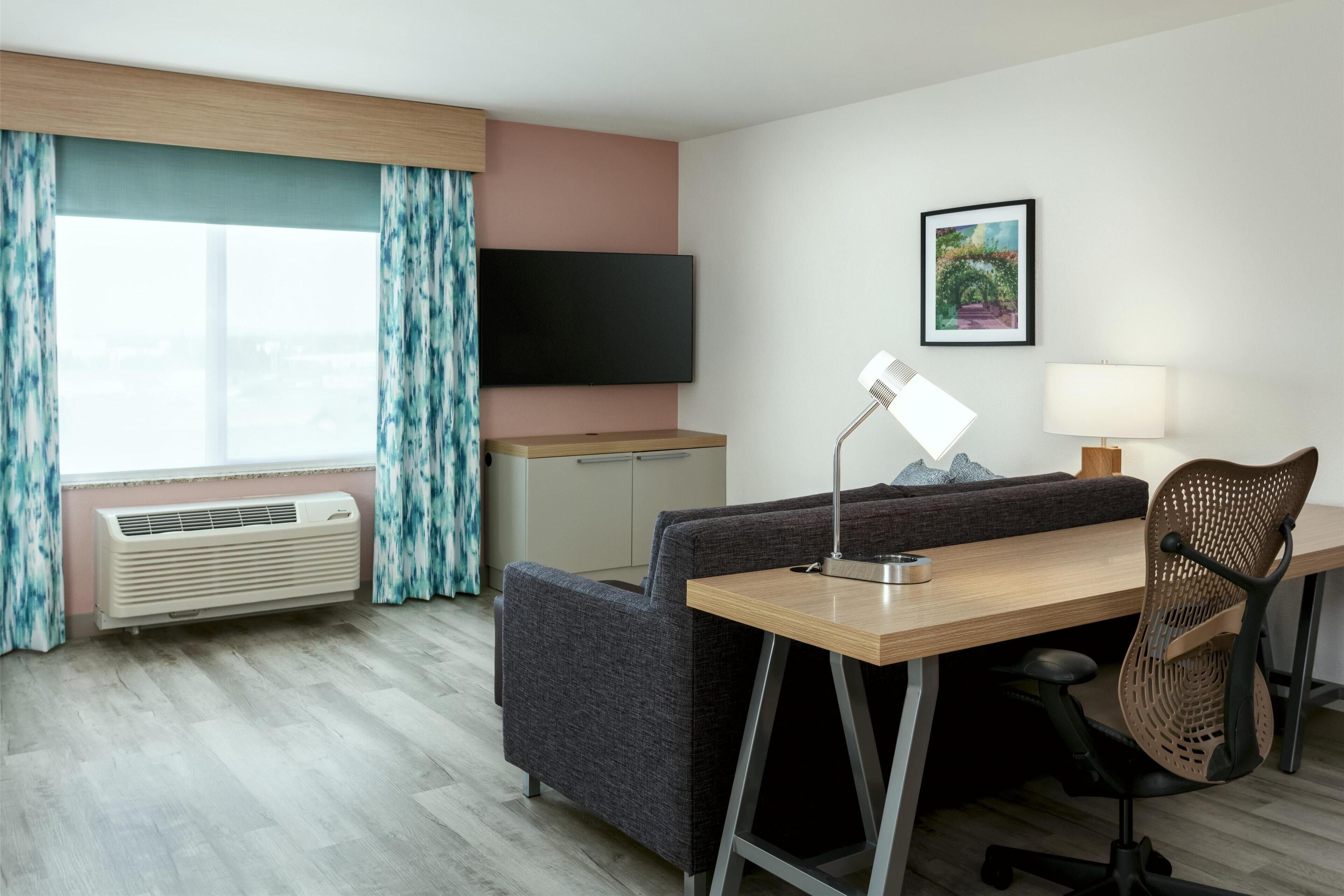 Une chambre d’hôtel équipée d’une télévision à écran plat, d’un bureau avec une chaise, d’un canapé, d’un lampadaire et d’une fenêtre avec des rideaux.