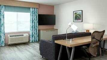 Une chambre d’hôtel équipée d’une télévision à écran plat, d’un bureau avec une chaise, d’un canapé, d’un lampadaire et d’une fenêtre avec des rideaux.