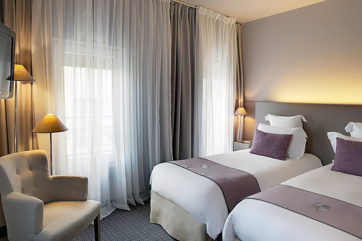 Ein Hotelzimmer mit zwei Betten, einem Sessel und einem großen Fenster mit durchsichtigen Vorhängen.